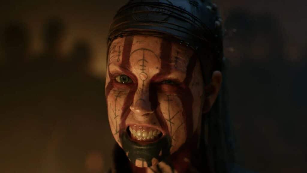 In a scene from Hellblade II, a person with face paint, blood-like marks, and an intense expression snarls at the camera in dim light—like a scene from one of the scariest games to play this halloween.