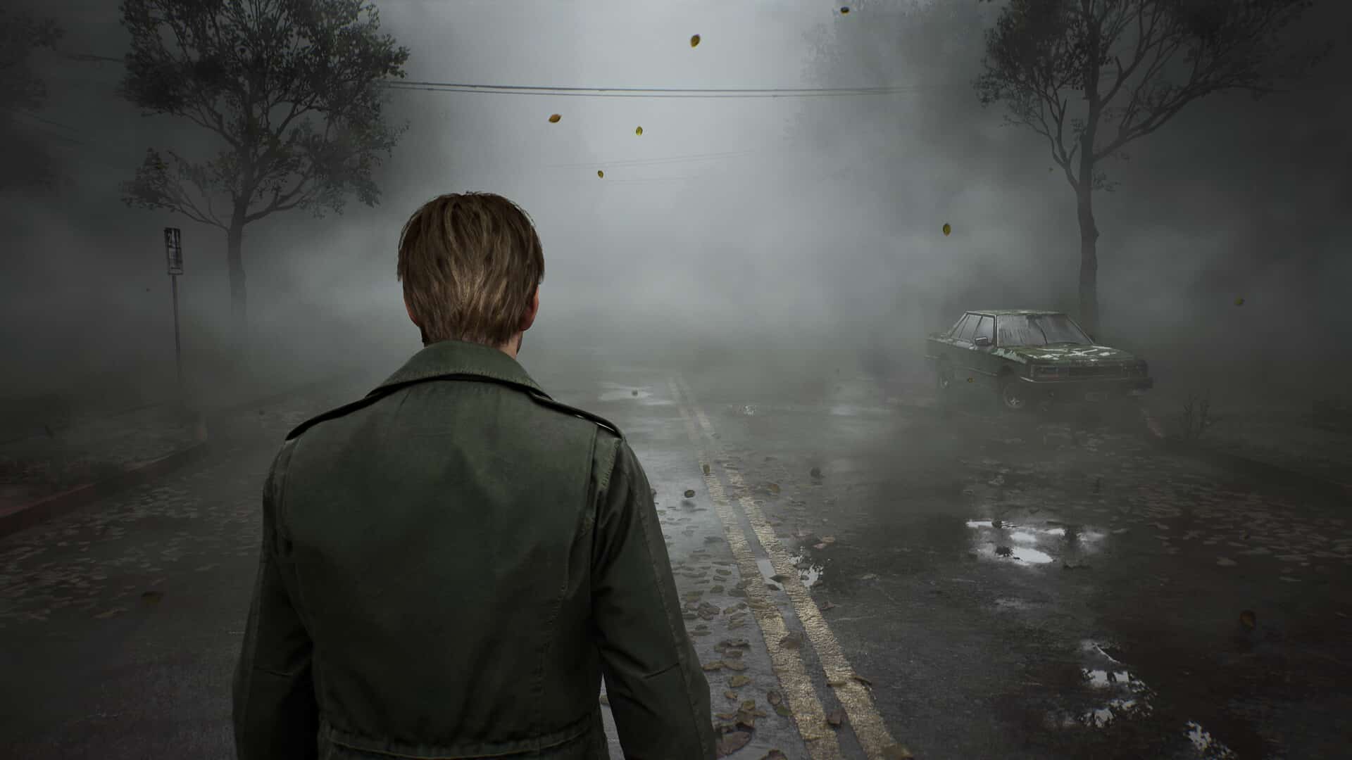 James Sunderland, in a green coat, stands on a foggy road, evoking the eerie atmosphere of Silent Hill 2 remake, with trees and an old car hauntingly silhouetted in the background.