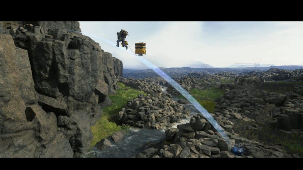 A person in a suit leaps with cargo over rocky terrain and a stream, guided by a blue line, reminiscent of the epic journeys found in Death Stranding Director’s Cut.