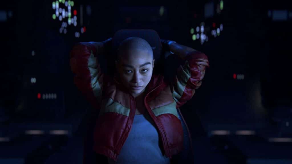 A person with a shaved head leans back in a dimly lit, hi-tech room, wearing a red and gold jacket in a scene from Intergalactic: The Heretic Prophet.