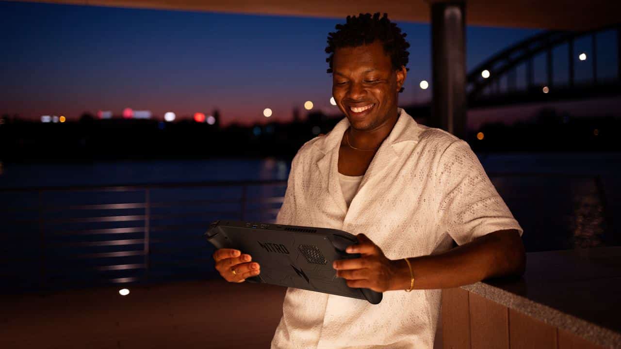 A person smiles while holding their Acer Nitro Blaze handheld gaming device outdoors at dusk, near a shimmering body of water.