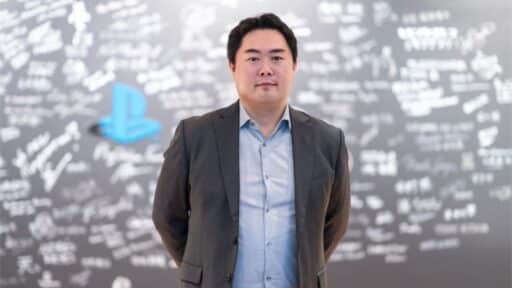 Hideaki Nishino, newly appointed CEO of Sony Interactive Entertainment, stands in a suit before a wall covered with signatures, accentuated by the iconic blue PlayStation logo.