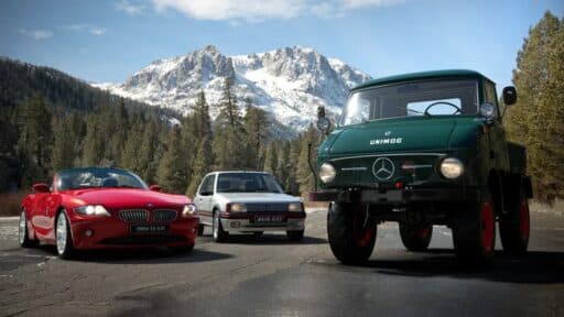 A red sports car, a white hatchback, and a green Unimog truck are parked on a mountain road with snowy peaks in the background, reminiscent of the stunning landscapes featured in Gran Turismo 7's update 1.56.