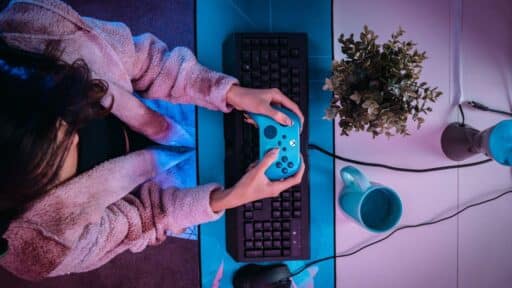 A person in a fur jacket is immersed in gaming, wielding a blue game controller over a keyboard and mouse on the desk, as if exploring the limitless possibilities of AI in gaming.