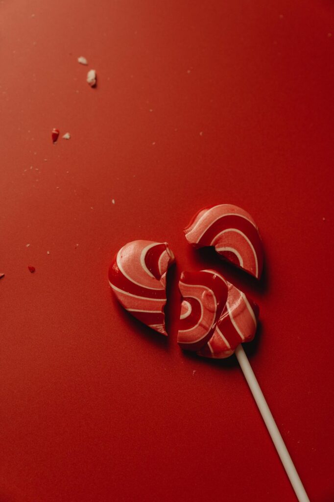 A shattered red and white heart-shaped lollipop rests on a vibrant red backdrop, surrounded by scattered candy pieces—perfect for setting the scene for Valentine's Day games for singles.