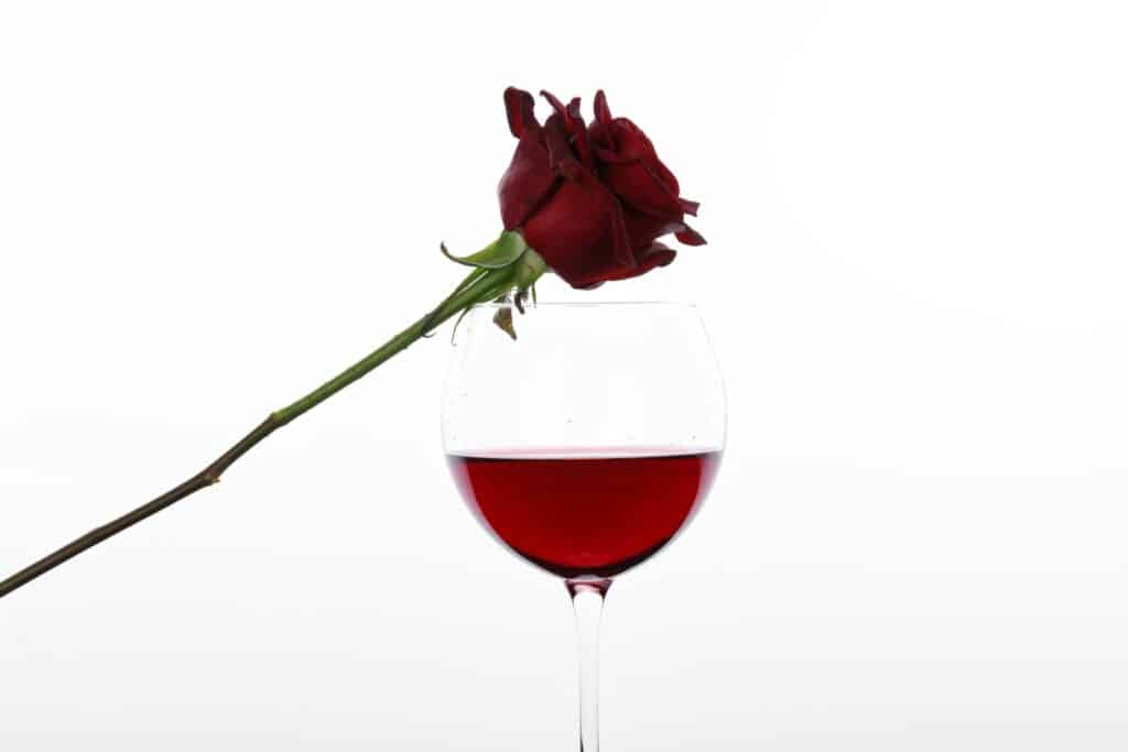 A red rose delicately rests on the rim of a wine glass filled with rich red liquid, set against a plain white background—perfect for staging unforgettable Valentine's Day games for singles seeking a touch of romance.