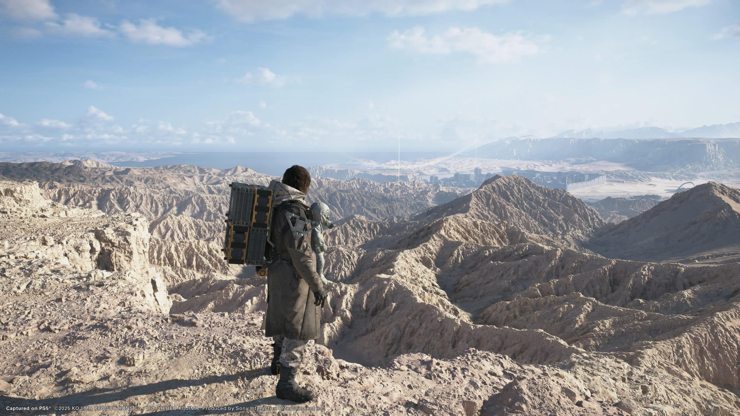 A lone figure stands on rocky terrain, contemplating the vast, rugged landscape under a clear blue sky, reminiscent of the surreal vistas from Death Stranding 2.