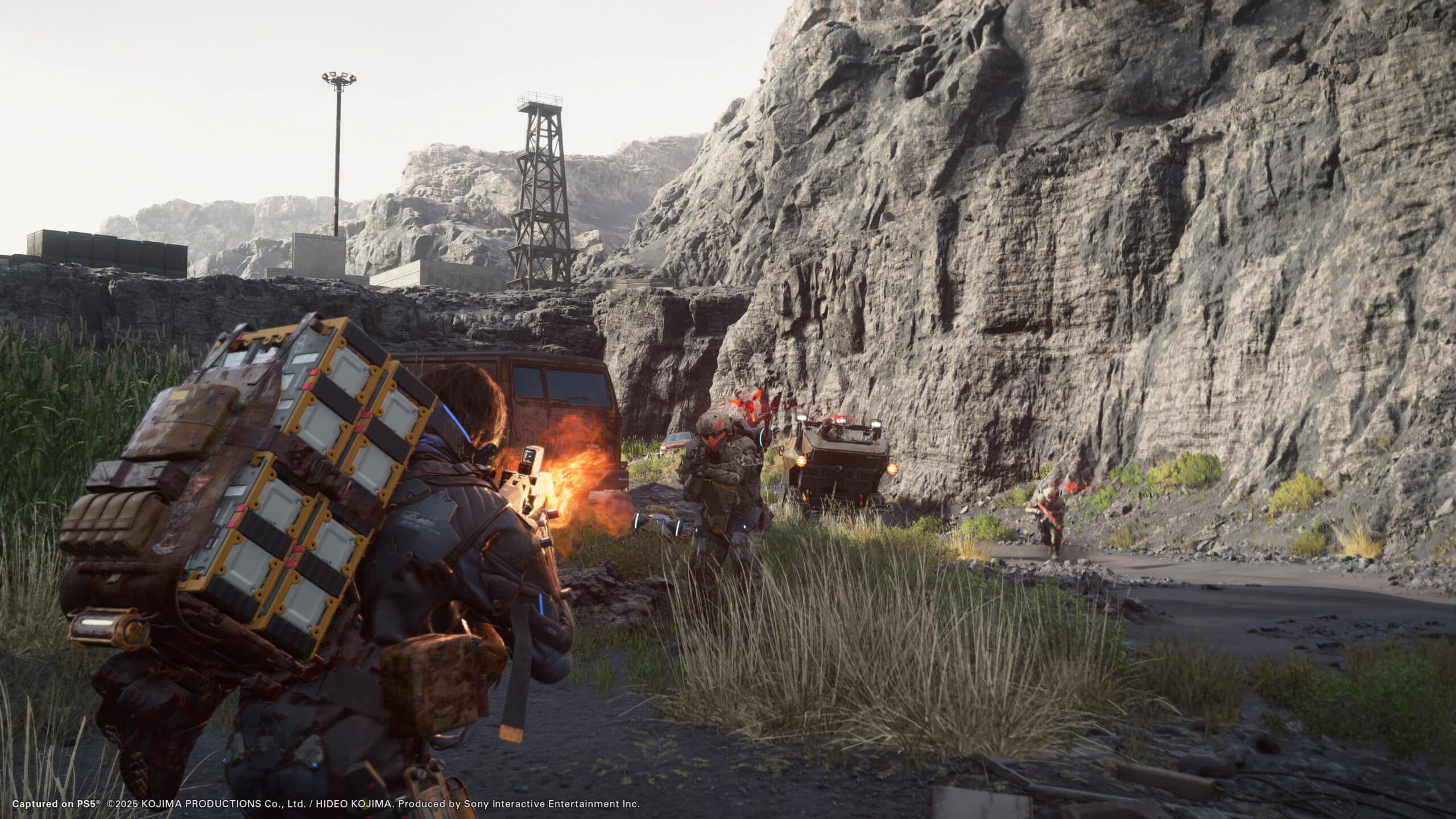 In a scene reminiscent of Death Stranding, a lone figure navigates rocky terrain, balancing packages amid looming industrial structures and parked vehicles, echoing the game's mysterious world.