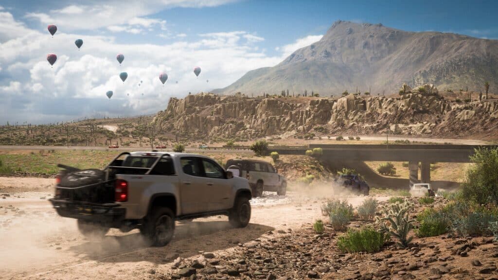 Pickup trucks race on a dusty desert road with mountains, hot air balloons, and an overpass in the background, capturing the thrilling spirit of Forza Horizon 5 PS5.