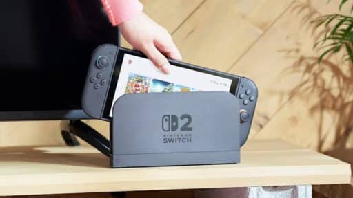 A hand places a Nintendo Switch OLED into its dock on a wooden TV stand next to a television, reminiscent of setups discussed in the recent Nintendo Switch 2 Ask the Developer interview.