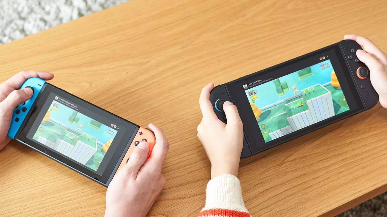 Two people enjoy Nintendo shared digital game access, playing the same video game on handheld consoles at a wooden table.