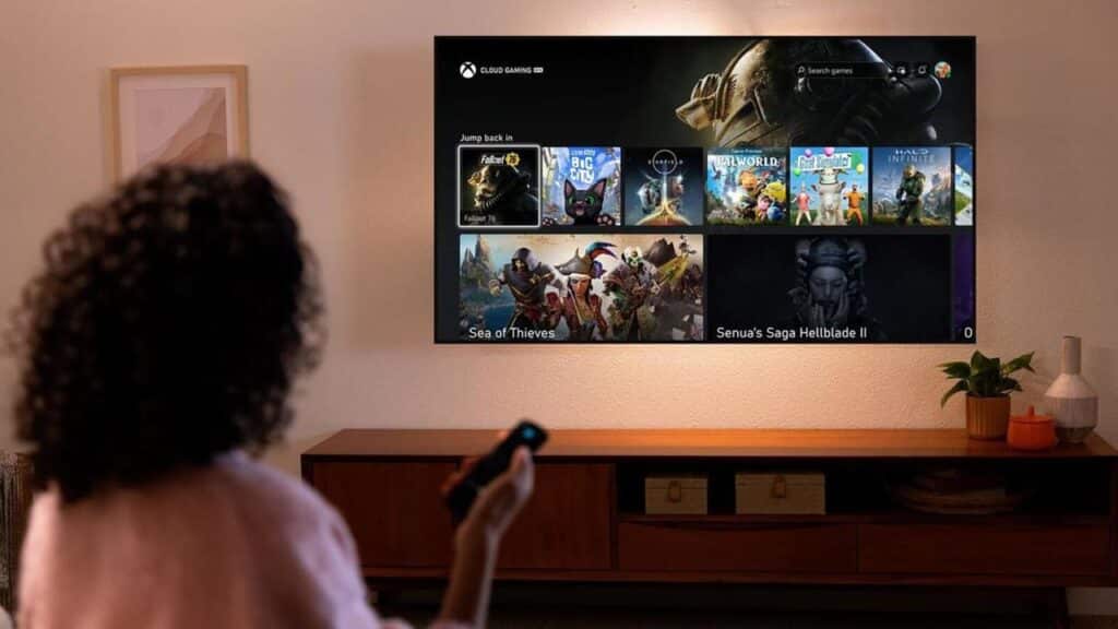 A person holds a remote and faces a wall-mounted TV displaying a video game selection screen, highlighting the impact of Xbox Q3 FY25 gaming revenue on popular choices.