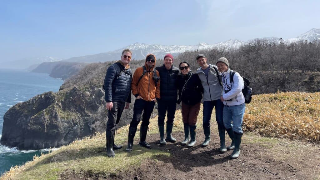Six people in outdoor gear stand on a cliffside overlooking mountains, sea, and dry grass, as the Ghost of Yotei looms mysteriously in the distant landscape.