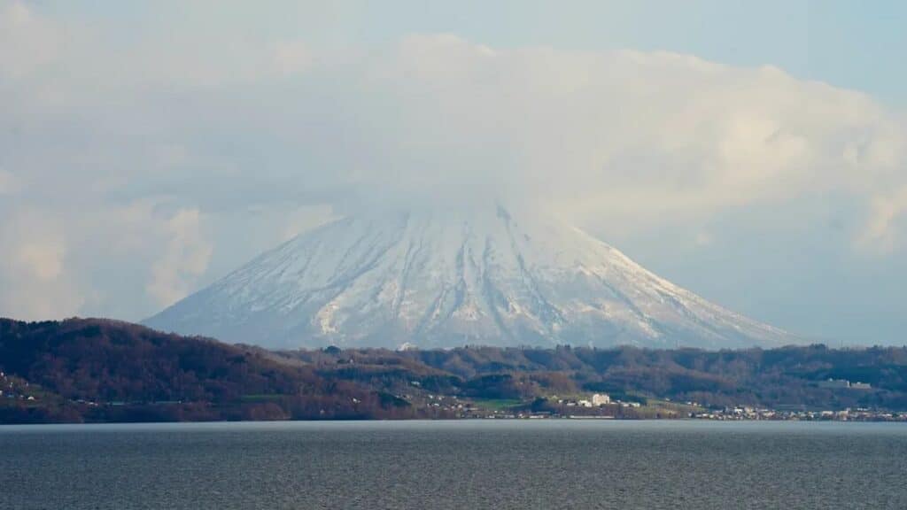 Snow-capped mountain partially covered by clouds—often called the Ghost of Yotei—rises above a tranquil lake and rolling hills in the foreground.