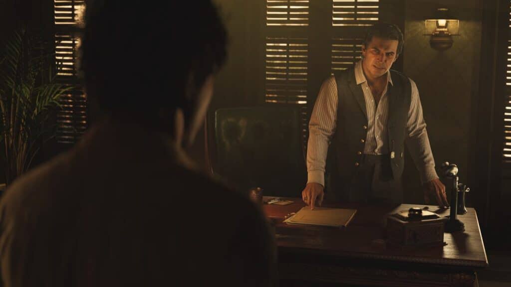 A man in a vest stands behind a desk, facing another person in a dimly lit office—reminiscent of scenes discussed at the Mafia: The Old Country PAX East panel.