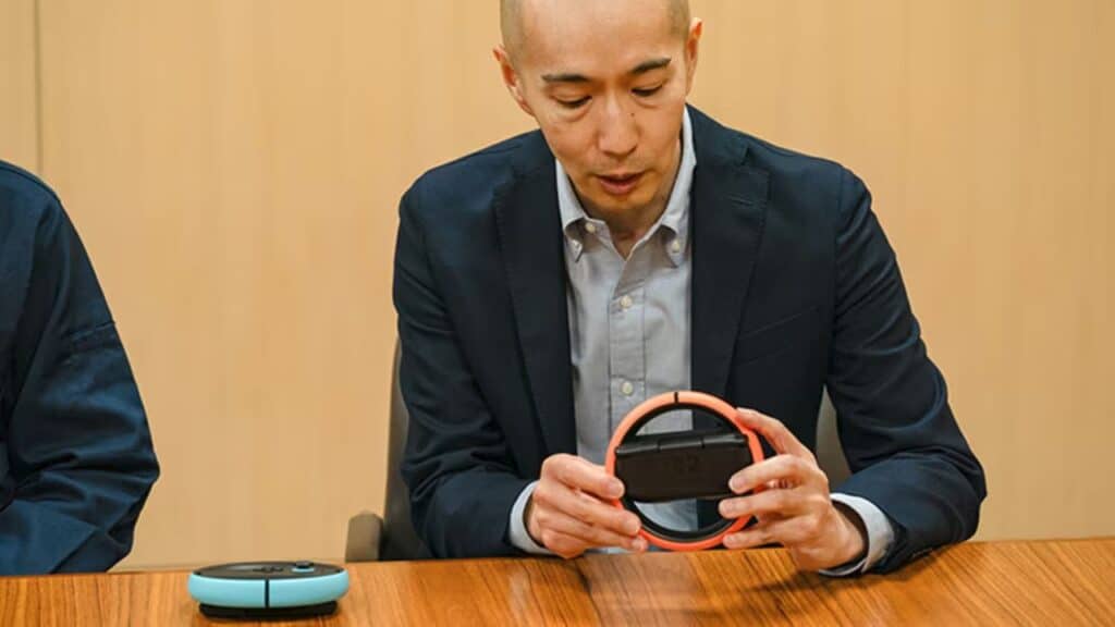 A man in a suit examines a red circular device inspired by Mario Kart World on Nintendo Switch 2 while seated at a wooden table.