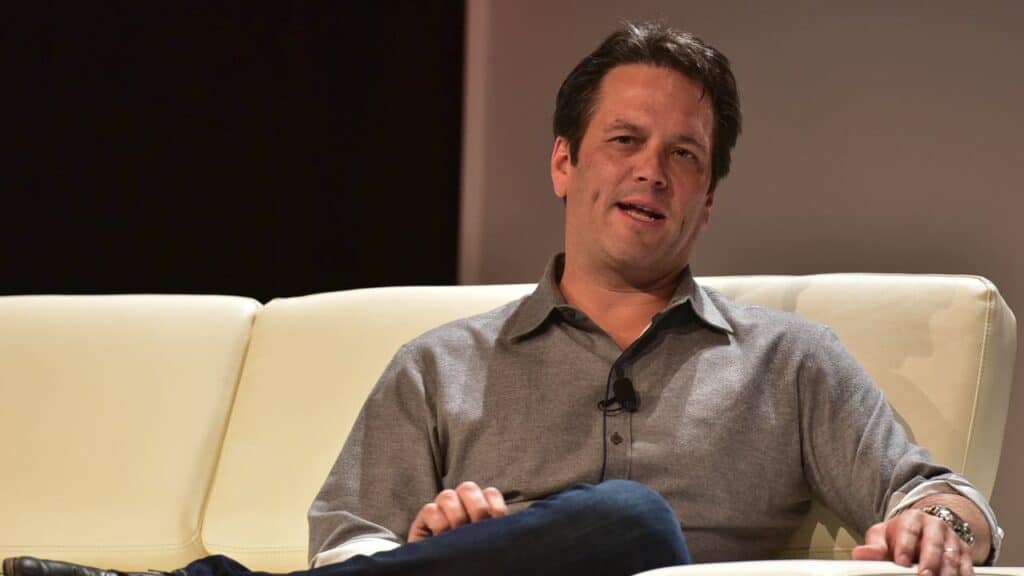 Phil Spencer in a gray shirt sits on a white couch, speaking and gesturing with his hand as he discusses the PlayStation engagement strategy.
