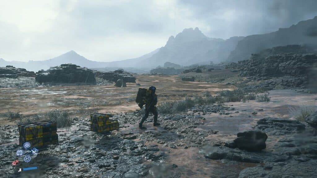 A person with a large backpack walks in a rocky, open landscape under a cloudy sky, with two boxes nearby—an image reminiscent of Death Stranding 2: On The Beach.