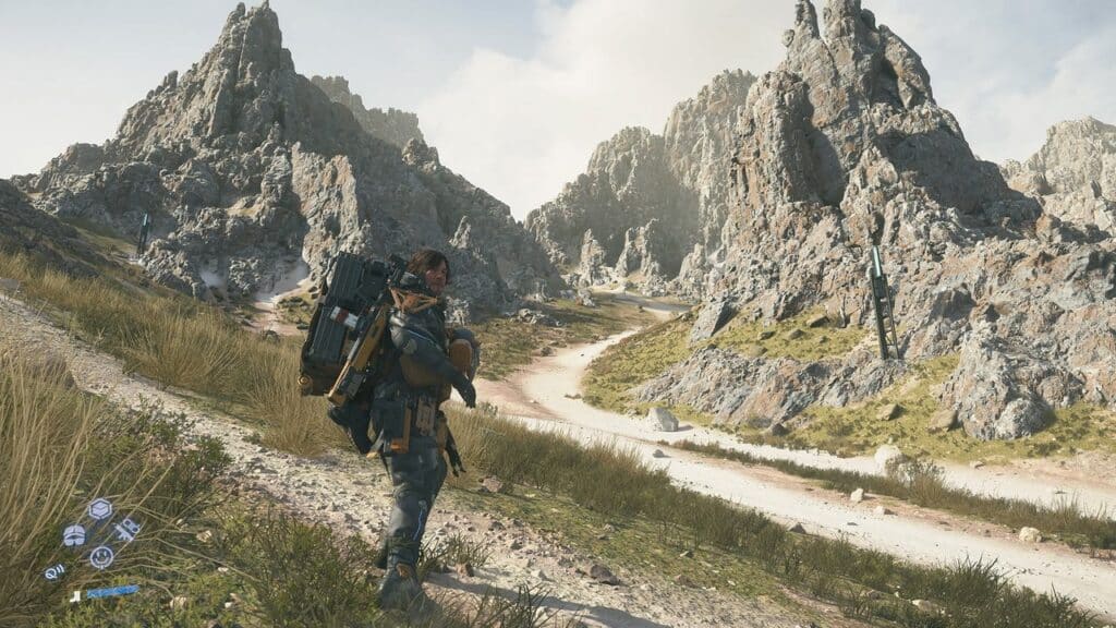 A person with heavy gear walks along a winding dirt path through rocky, mountainous terrain under a cloudy sky, evoking scenes from Death Stranding 2: On The Beach.