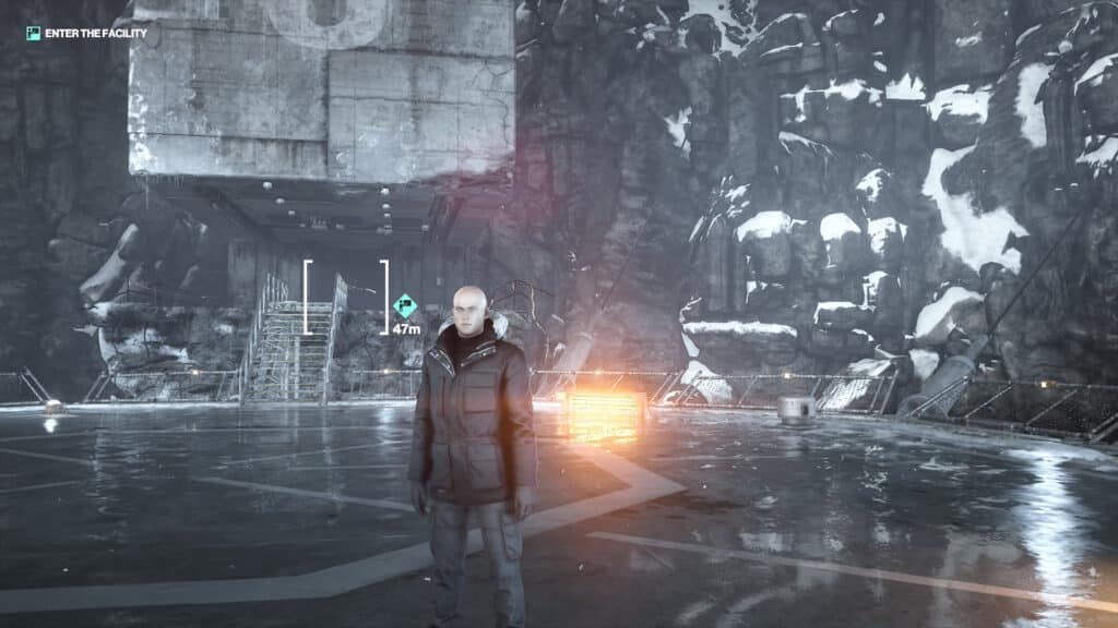 A bald man in a dark coat stands outside a snowy, industrial facility with icy mountains behind him, evoking the chilling atmosphere of Hitman World of Assassination Signature Edition.