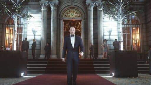 A bald man in a tuxedo walks down a red carpet outside a grand, ornate building at night, reminiscent of an IO Interactive interview scene.