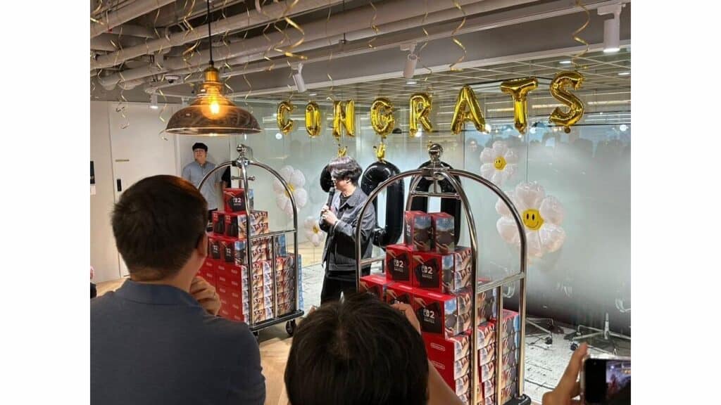 A person holds a microphone near "CONGRATS" balloons, with stacks of gifts, people taking photos, and a Stellar Blade Nintendo Switch 2 nestled among the presents.