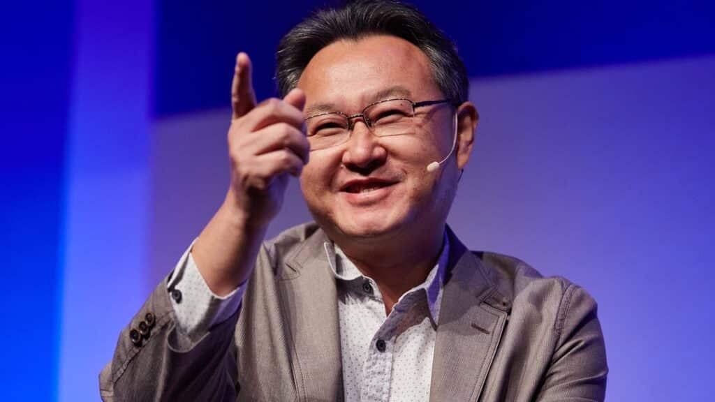 Smiling man in a suit jacket gestures and points, wearing glasses and a microphone headset on stage as Shuhei Yoshida talks Nintendo.
