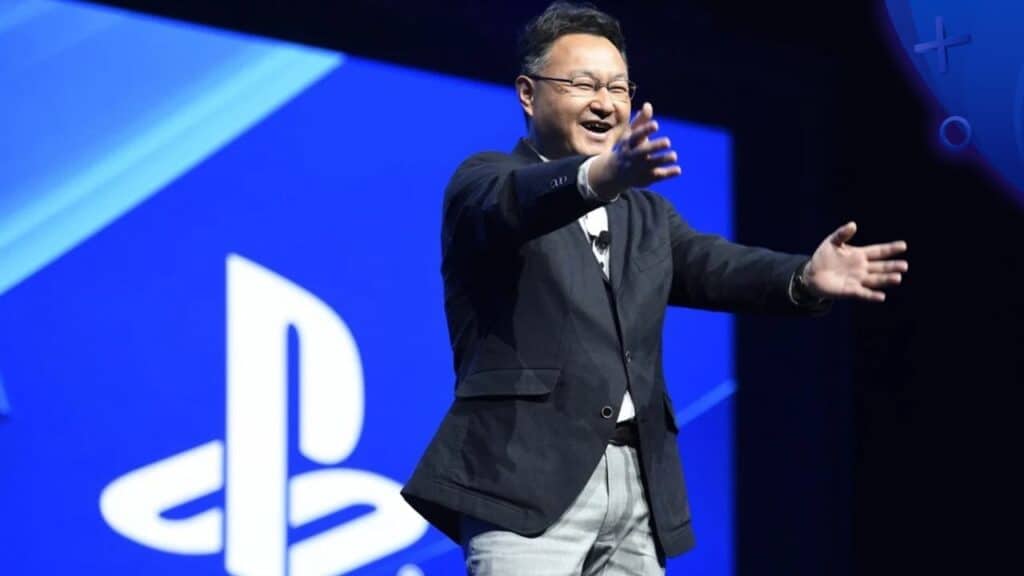 A man in a suit smiles with open arms on stage in front of a large PlayStation logo, as Shuhei Yoshida talks Nintendo and celebrates gaming unity.