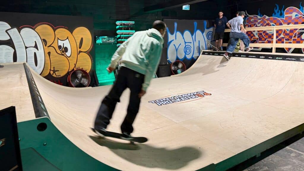 Three people skateboard on an indoor ramp at a spot reminiscent of a Tony Hawk’s Pro Skater 3 + 4 Skate Hang, with colorful graffiti decorating the walls in the background.