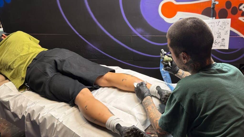 Tattoo artist working on the back of a client’s leg in a tattoo studio with dark, colorful walls, giving off serious Tony Hawk’s Pro Skater 3 + 4 Skate Hang vibes.