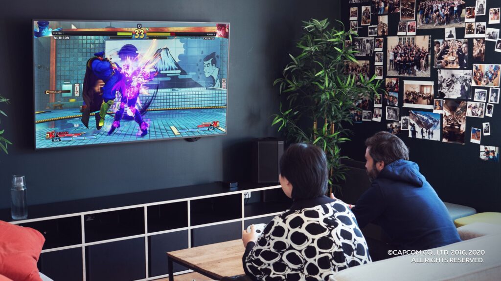 Two people play a fighting video game on a wall-mounted TV in a cozy room with photos and plants.