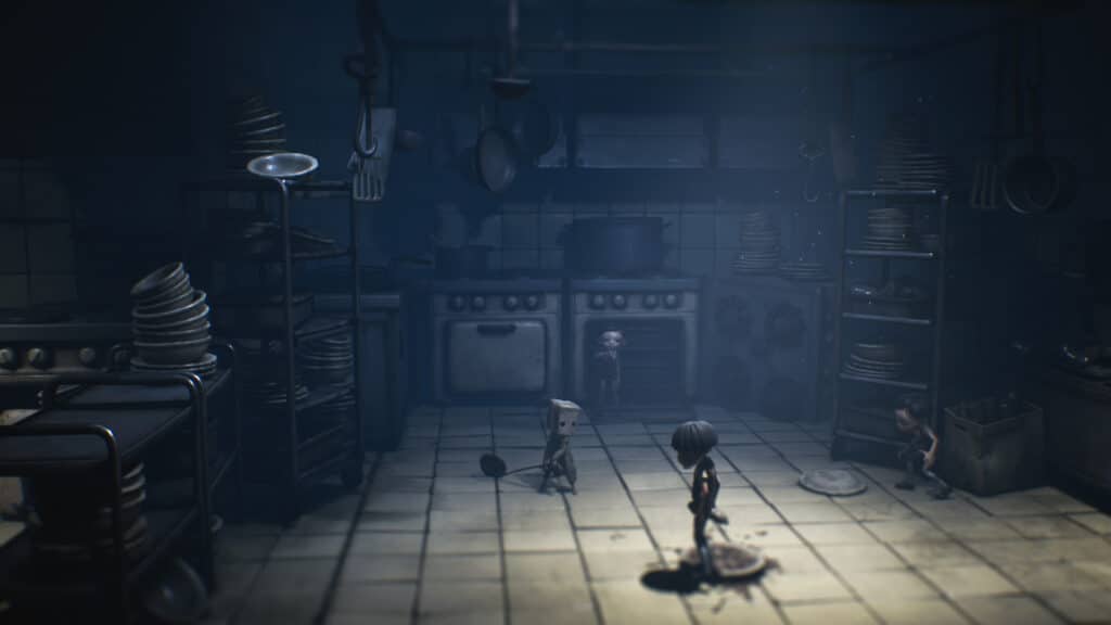 Two small characters stand in a dark, cluttered kitchen filled with dirty dishes and large cooking utensils, evoking the eerie atmosphere of Little Nightmares II.