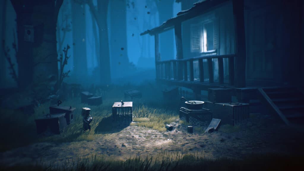 A dimly lit cabin in a foggy forest, reminiscent of Little Nightmares II, stands with scattered cages and wooden debris strewn across the yard at night.