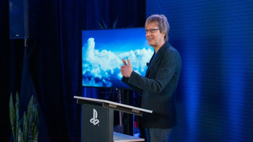Mark Cerny gives a thumbs up while speaking at a PlayStation podium, with a screen of clouds behind them, possibly hinting at the upcoming PS5 Pro PSSR upgrade.