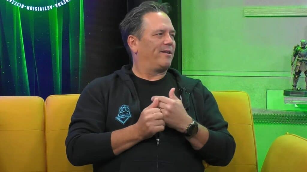 Phil Spencer sits on a yellow couch, gesturing with his hands, in a green-lit room as news of Xbox layoffs trends in the background.