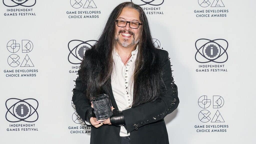 A man with long hair and glasses smiles, holding an award at a Game Developers Choice Awards event, celebrating creativity in a year marked by Xbox layoffs.