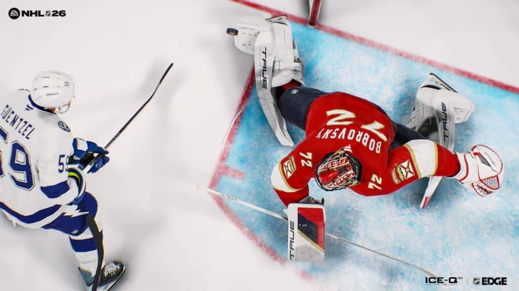 A hockey goalie in red makes a save as a player in white approaches the goal, viewed from above—capturing the action-packed excitement of EA Sports NHL 26.