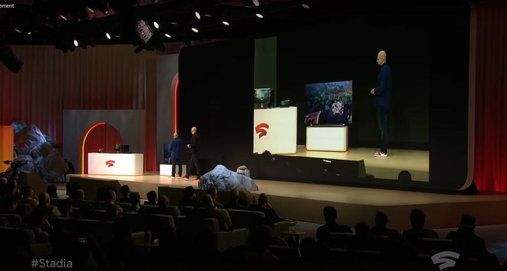 A presenter on stage demonstrates Stadia gaming to an audience, with gameplay shown on a large screen during the Google Stadia GDC 2019 announcement.