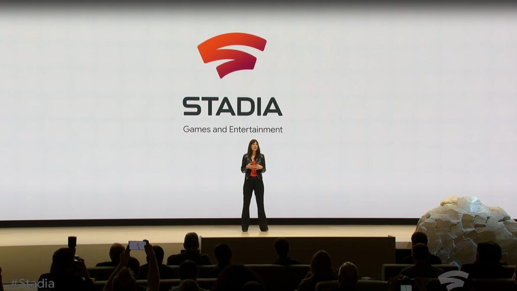 Jade Raymond stands on stage presenting Stadia, with its logo and "Games and Entertainment" on a large screen behind her, marking the Google Stadia shutdown announcement anniversary.