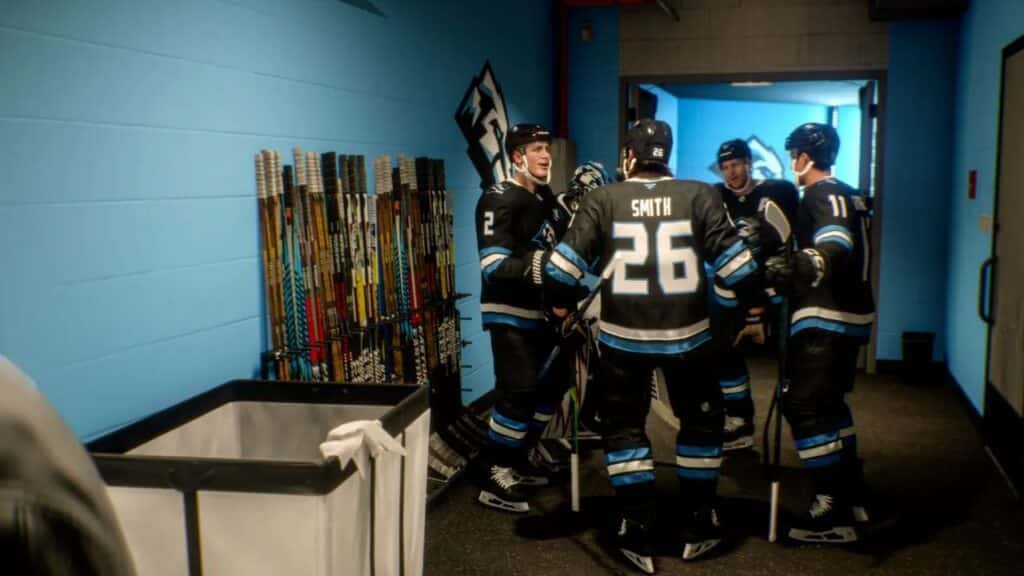 Five hockey players in black uniforms talk in a hallway near a rack of hockey sticks and a laundry bin in NHL 26.