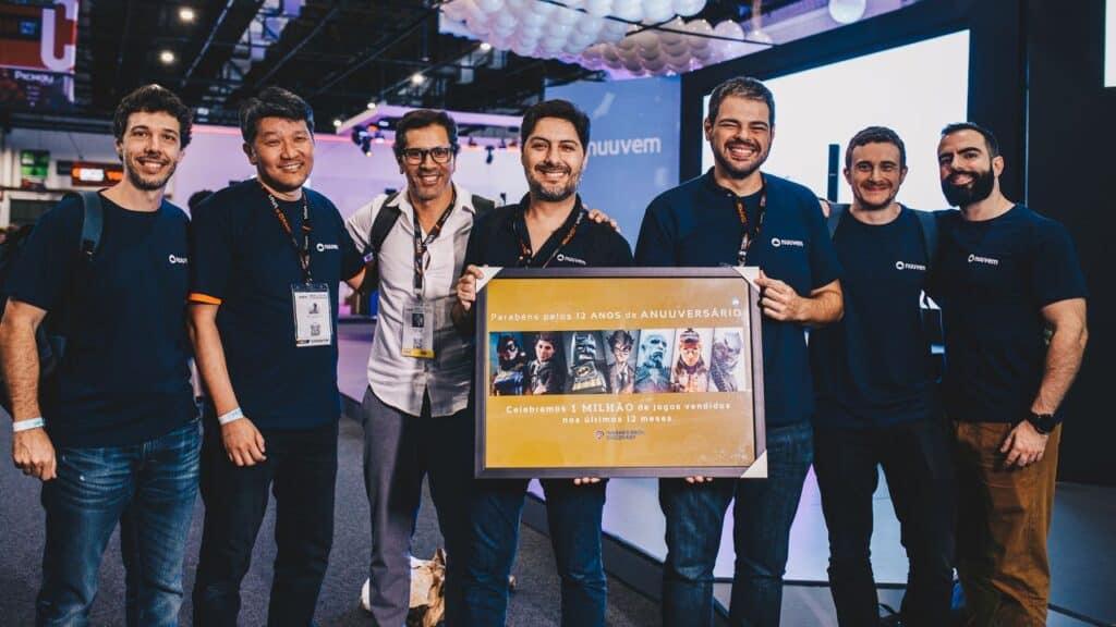 Fernando Campos smiling with team stand ing together indoors, holding a large anniversary sign with images and text, celebrating after a recent Nuuvem spawnd interview.