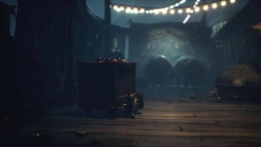 A dark carnival scene from Little Nightmares III, with a mysterious box, scattered apples, string lights, and large animals looming in the background.
