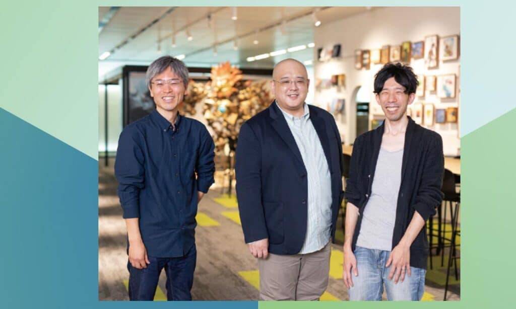 Three smiling men standing together in a modern, brightly lit office space with art on the walls, discussing PlayStation Network and AWS solutions.