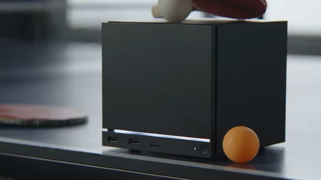 A small black cube-shaped device on a table, with ping pong paddles and an orange ping pong ball nearby.