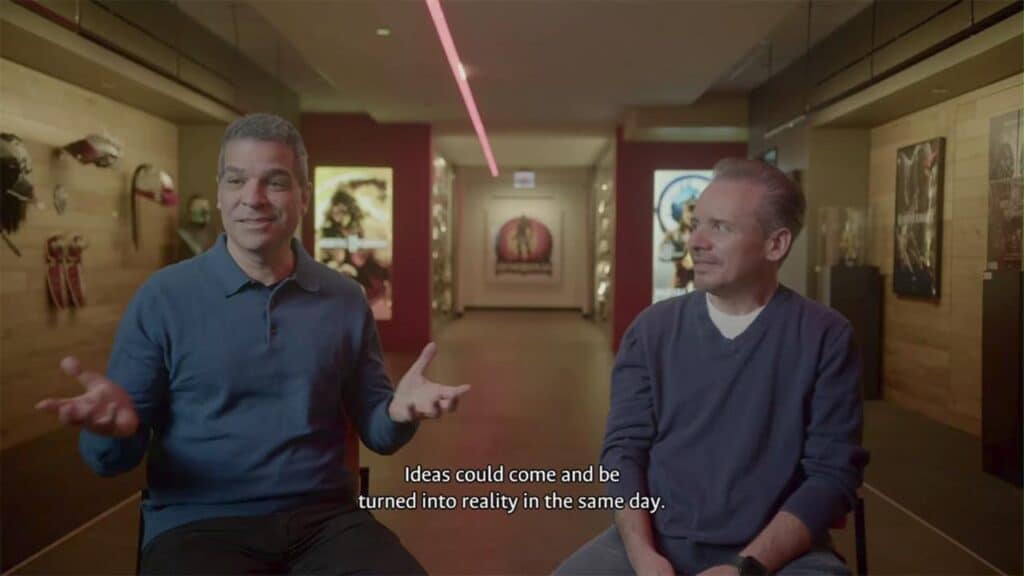 Two men sit in a hallway talking, with Mortal Kombat: Legacy Kollection movie posters and costumes on the walls; subtitles appear onscreen.