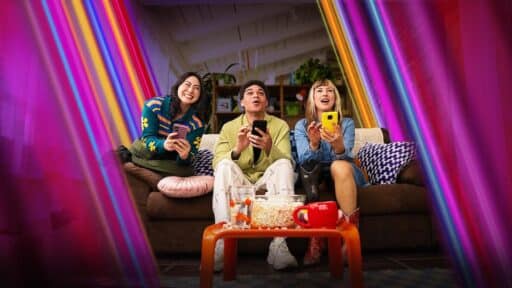 Three people sit on a couch, smiling and playing Netflix Games on TV on their phones, with popcorn and drinks on the table.