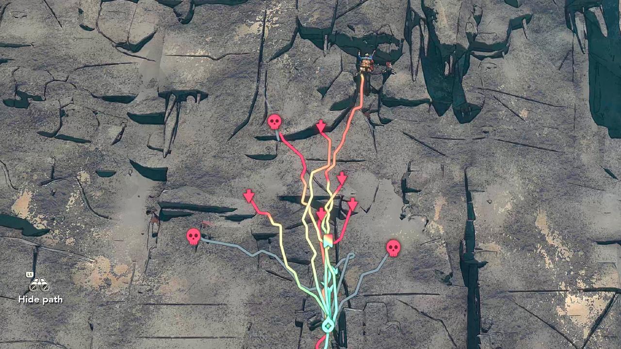 A rocky cliff face with six colorful climbing paths marked, ending with stars and skull icons, and a small cairn at the base to mark the trailhead in Cairn.