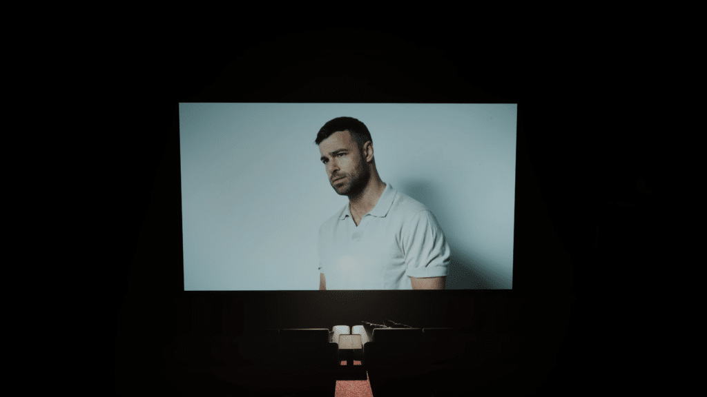 A close-up of a male actor's face displayed on a large screen in a dimly lit setting, evoking a cinematic atmosphere.
