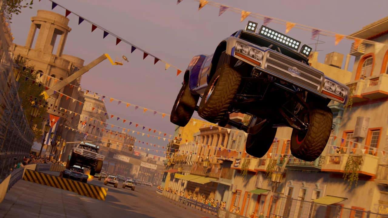 A racing truck from GRID Legends: Deluxe Edition soars over a ramp on a city street lined with colorful buildings and festive banners.