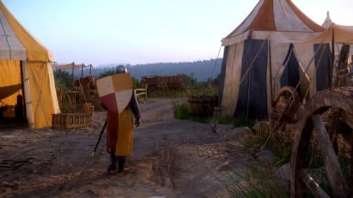 A knight with a shield walks near medieval tents and wooden carts on a dirt path at sunset.